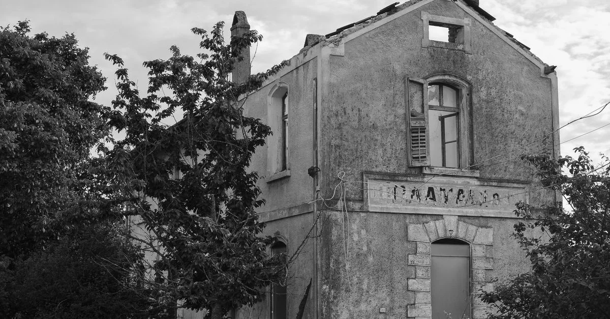 Photo en noir et blanc d'une maison rurale abandonnée, montrant la décomposition et la reprise par la nature.