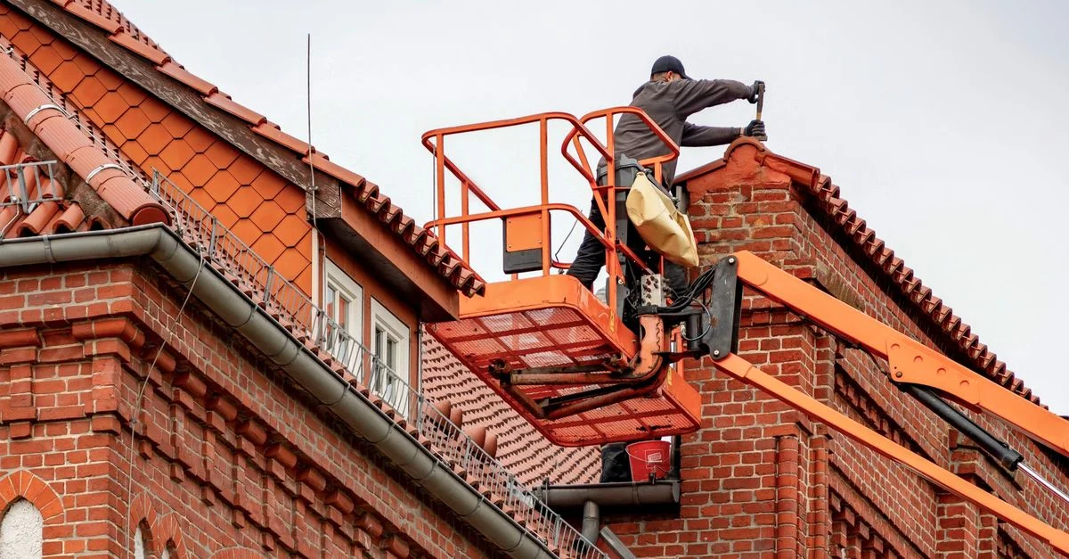 Couvreur dans une nacelle effectuant l'entretien d'un bâtiment en brique rouge avec un toit en tuiles.