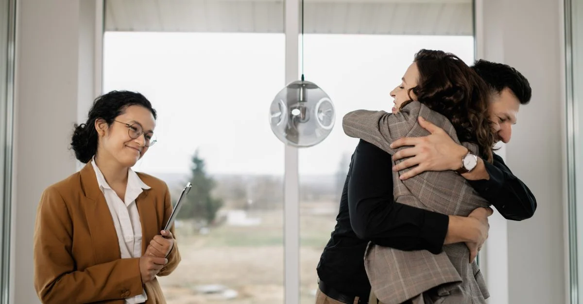 Un couple s'embrasse joyeusement dans leur nouvelle maison tandis qu'un agent immobilier satisfait les observe.